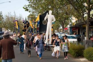 A group of people walking in an outdoor procession while carrying a white sculpture of a tall person 