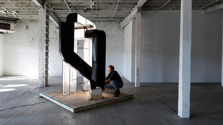 Figure bending on a platform while studying and touching an abstract sculpture in an industrial space.