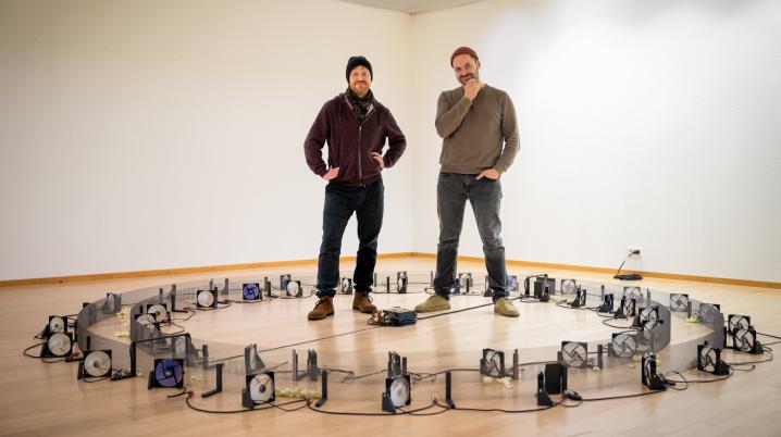 Two people in the midst of their art installations in an exhibition room, looking at the camera